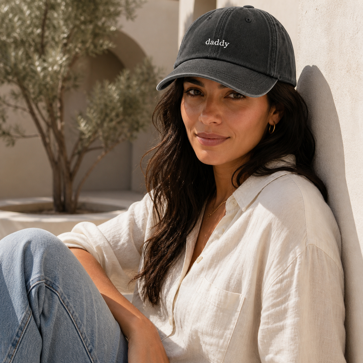 A woman wearing a dark charcoal gray baseball cap with the word 'daddy' embroidered in white on the front, sitting against a light-colored wall in a sunlit outdoor setting.