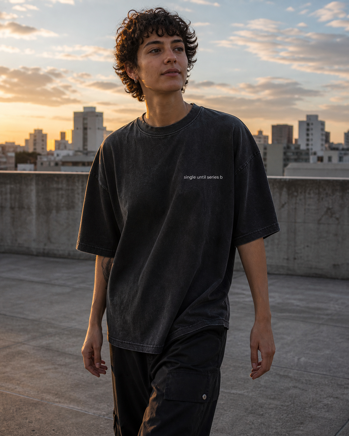 A person standing on a rooftop at sunset wearing an oversized faded black graphic T-shirt with small white text on the chest and dark cargo pants.