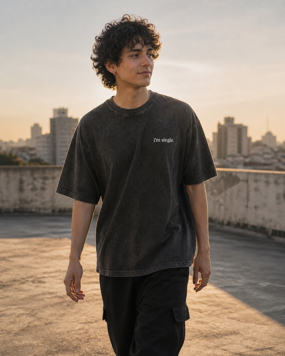 A man with curly dark hair stands on a rooftop at sunset wearing a faded black washed t-shirt with the text 'I'm single.' printed in white on the left chest, paired with black cargo pants.
