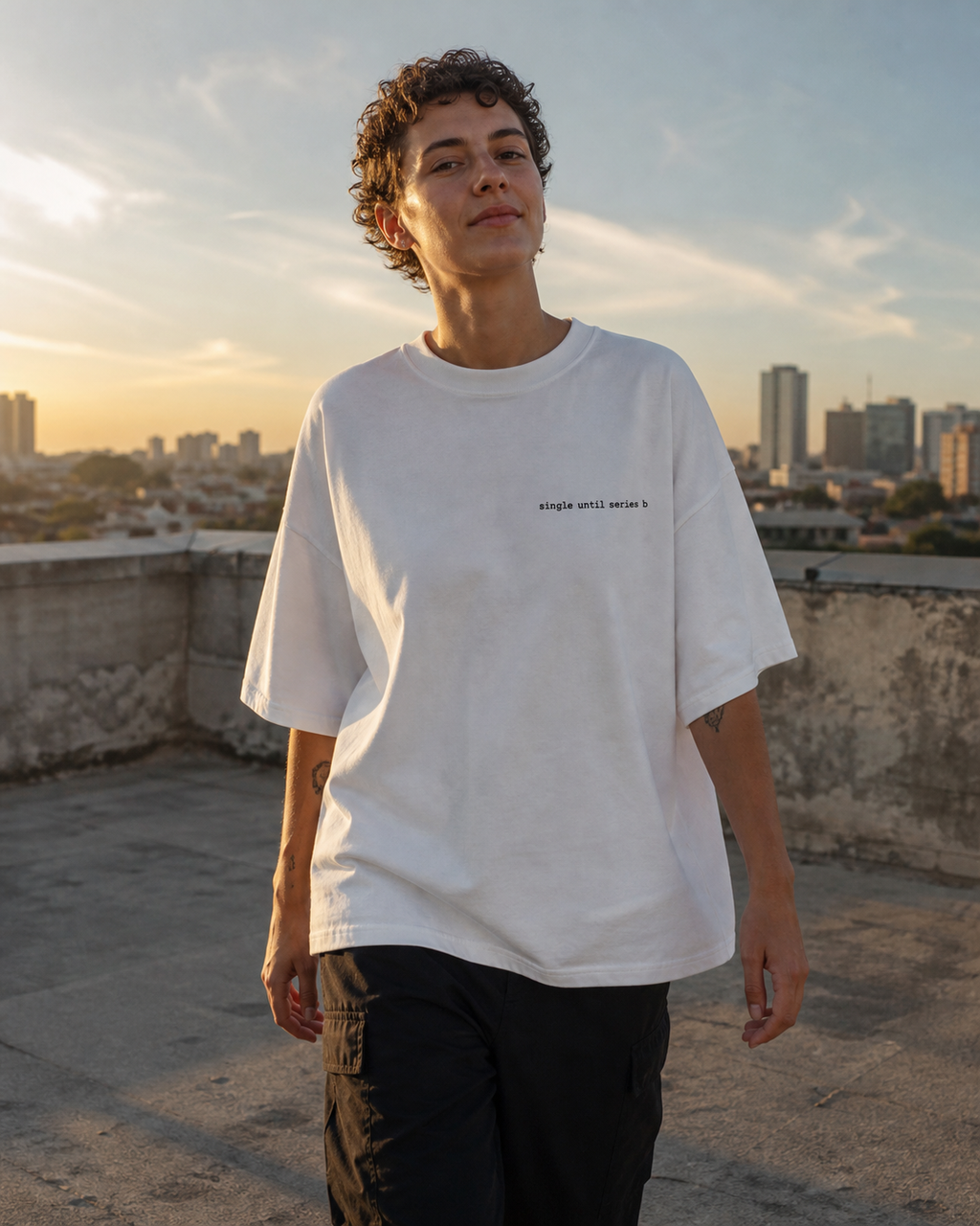 A person standing on a rooftop at sunset wearing an oversized white graphic t-shirt with small black text on the chest and dark cargo trousers.