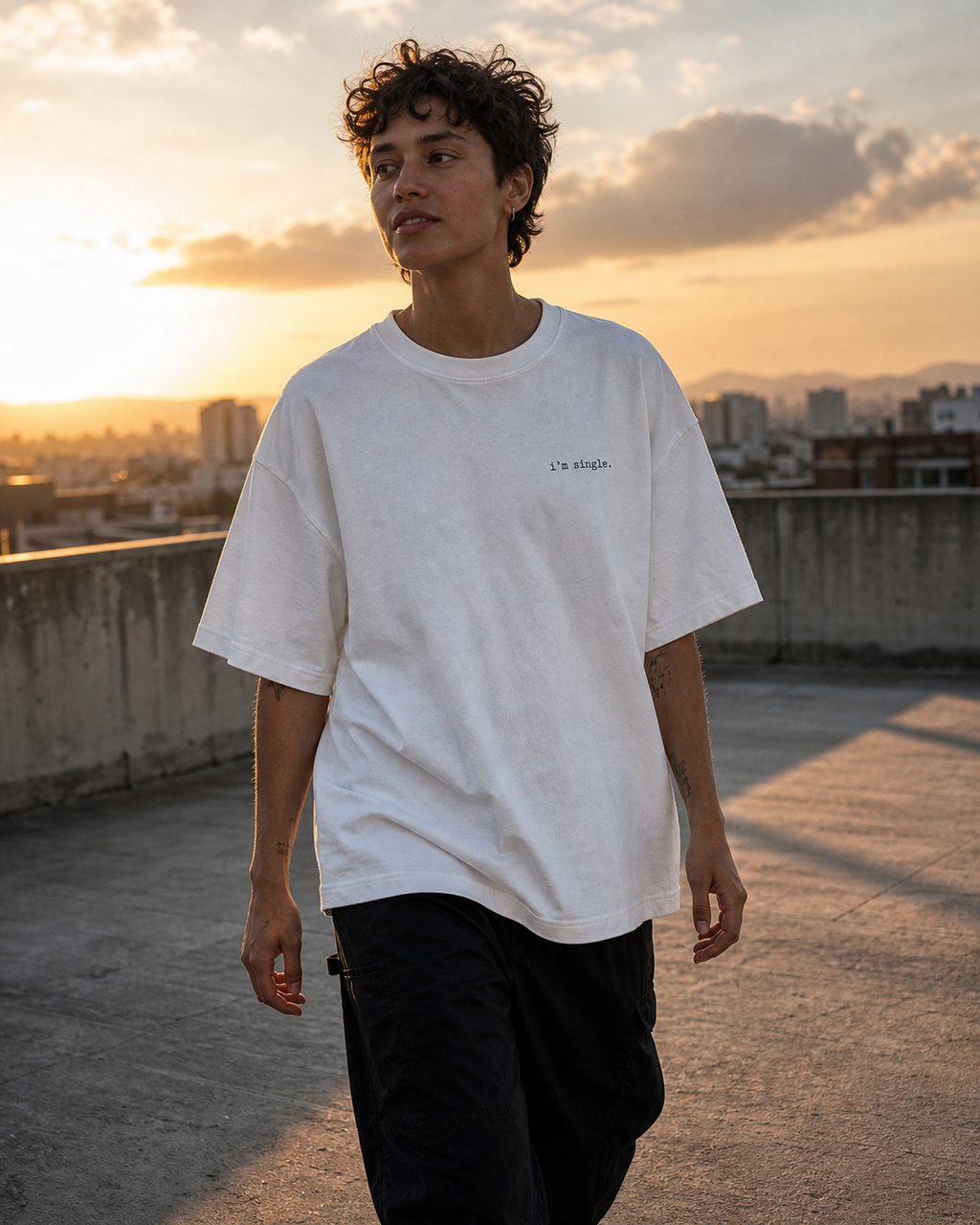 A man standing on a rooftop at sunset wearing a white t-shirt with 'i'm single.' printed in black on the left chest, paired with black cargo trousers.