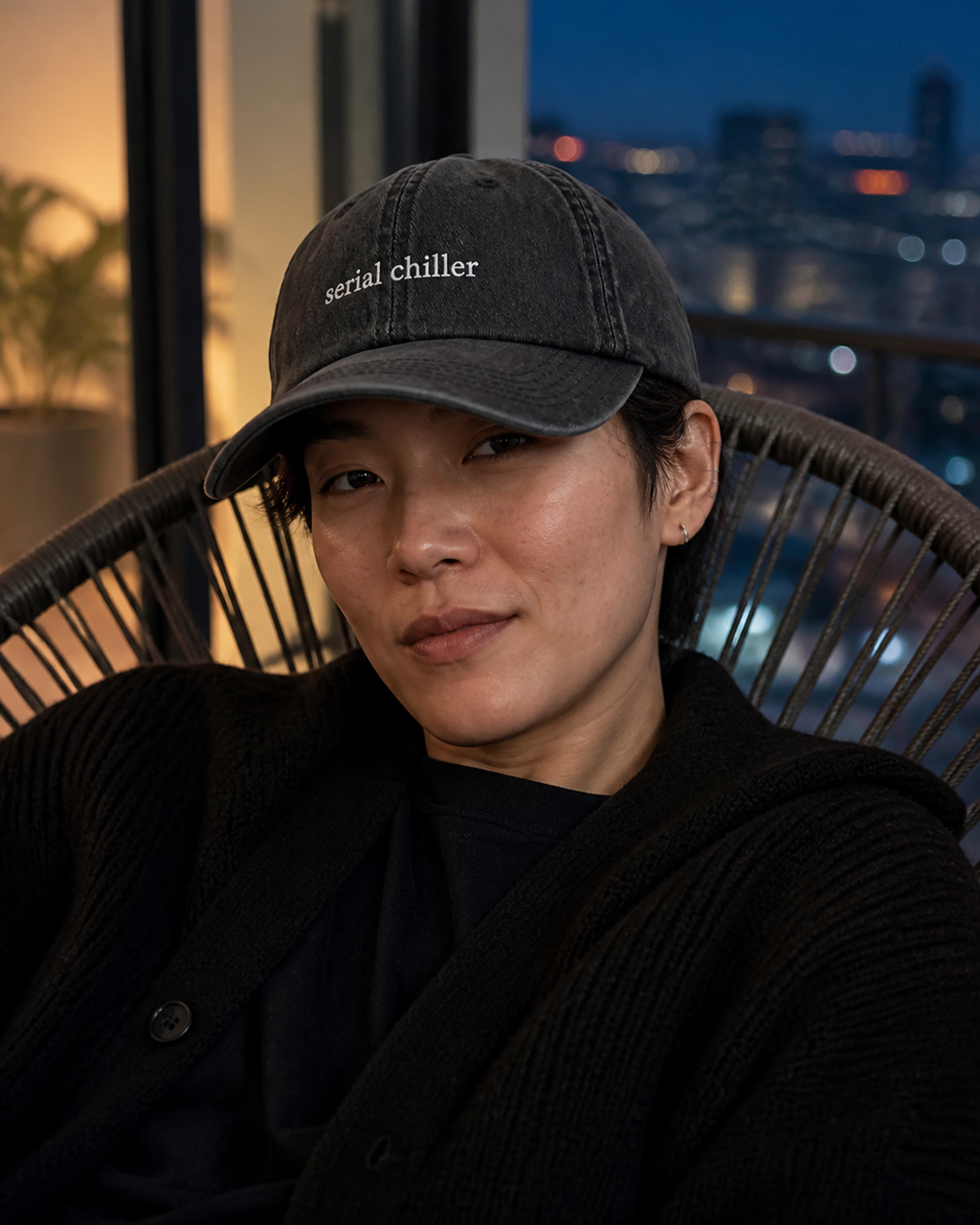 A person wearing a dark gray baseball cap with the embroidered text 'serial chiller' in white, seated in a wicker chair with a blurred city skyline at dusk in the background.