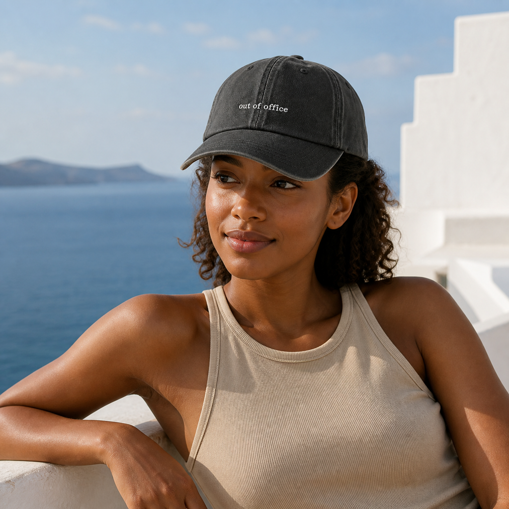 A woman wearing a dark charcoal gray baseball cap with 'out of office' embroidered in white on the front, paired with a beige ribbed tank top, posing outdoors with a blue sea and white buildings in the background.