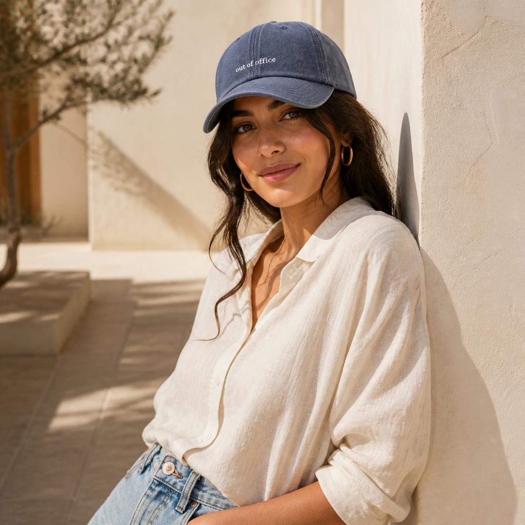 A woman wearing a blue baseball cap with 'rest of the office' embroidered on the front, a cream linen shirt, and light-wash jeans, leaning against a sunlit beige stucco wall.