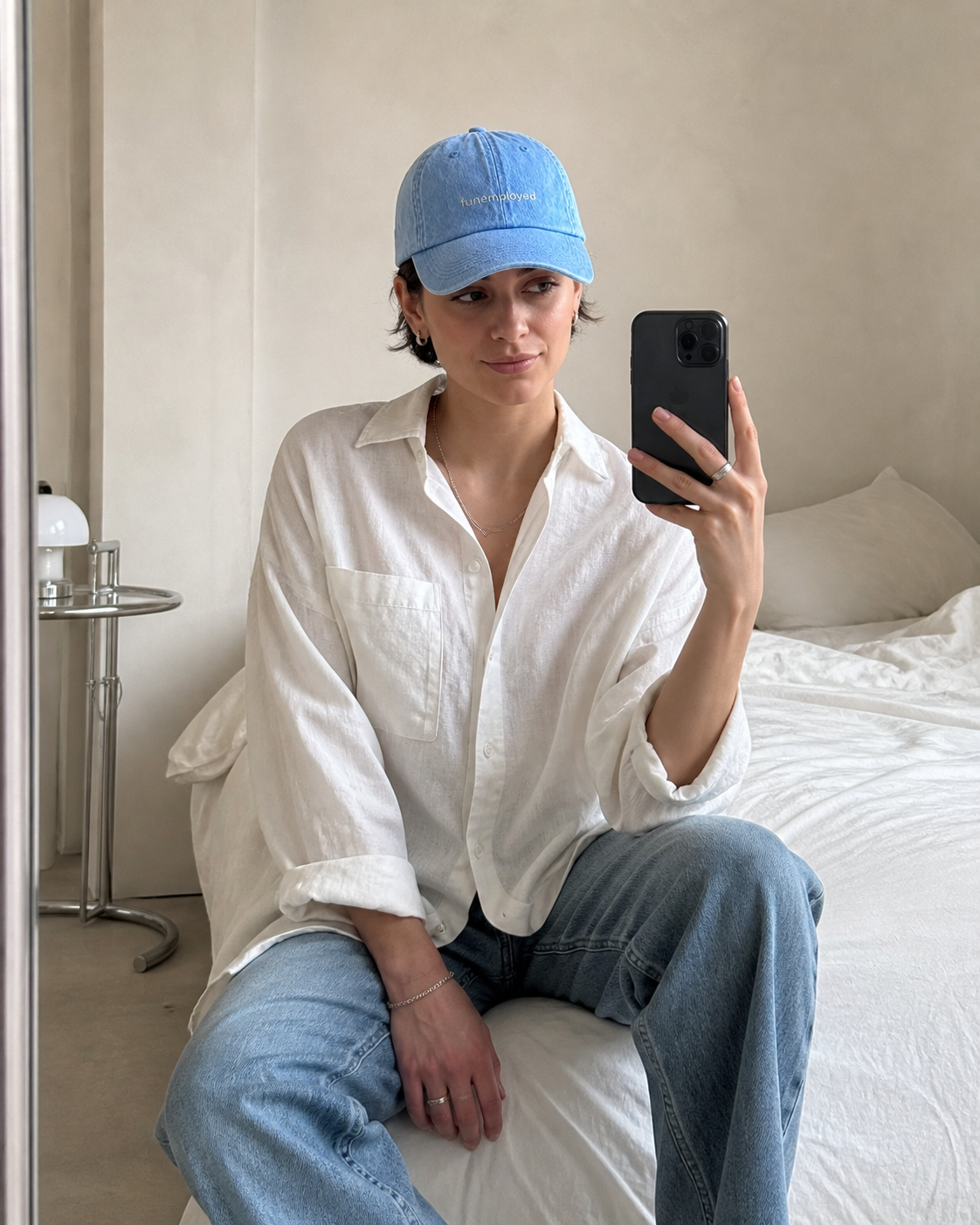 A person taking a mirror selfie in a bedroom, wearing a light blue embroidered baseball cap, a loose white button-up shirt, and light blue jeans.