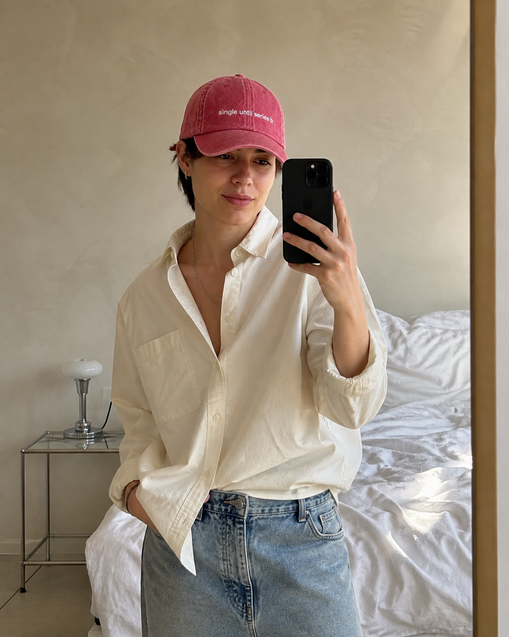 A person taking a mirror selfie in a bedroom, wearing a pink baseball cap with white embroidered text, an off-white button-down shirt, and light-wash blue jeans.