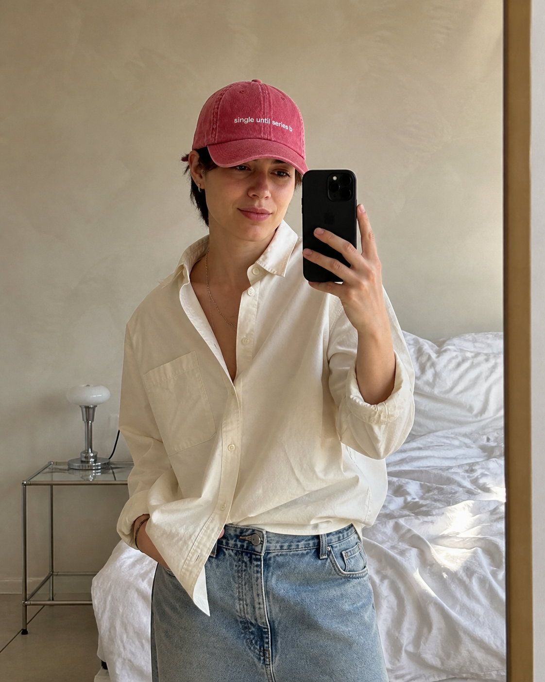 A person taking a mirror selfie in a bedroom, wearing a pink baseball cap with white embroidered text, an off-white button-down shirt, and light-wash blue jeans.