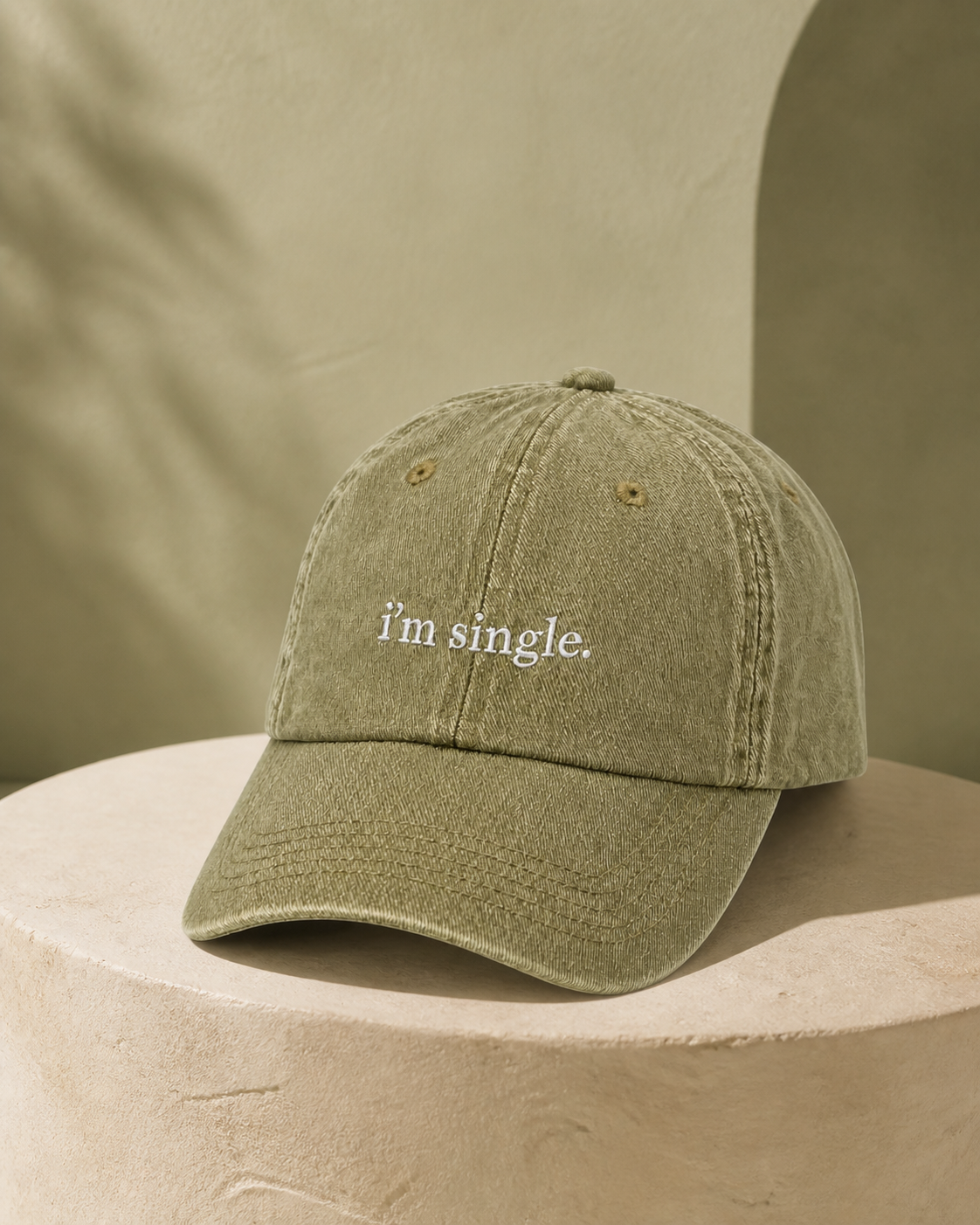 An olive green washed cotton baseball cap with the phrase 'i'm single.' embroidered in white on the front, displayed on a textured beige stone pedestal.