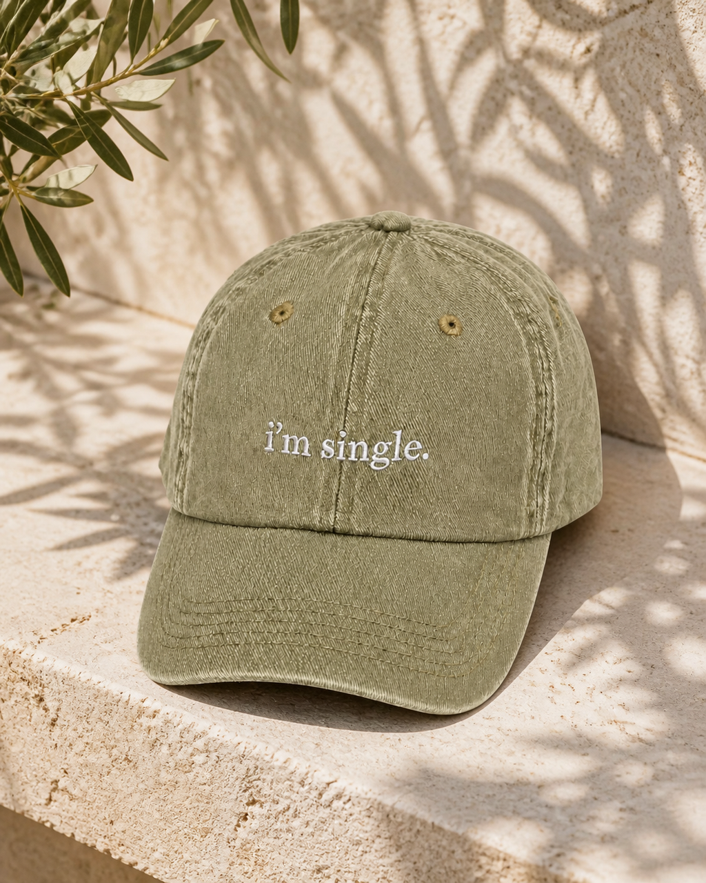 An olive green washed cotton baseball cap with the phrase 'i'm single.' embroidered in white script on the front, resting on a textured stone ledge with leaf shadows cast over it.