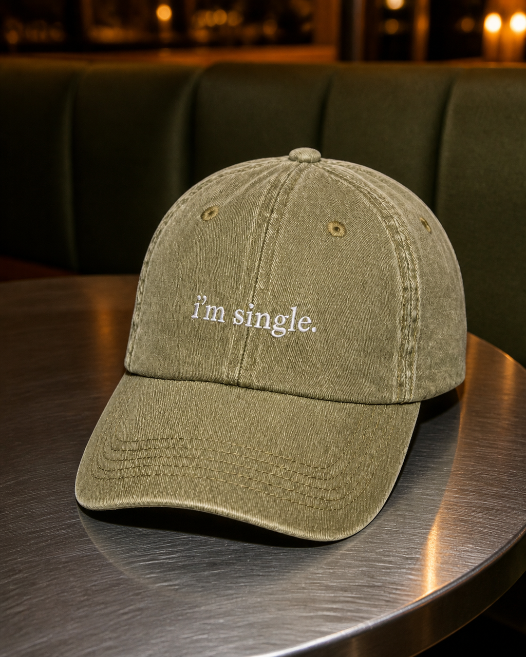 An olive green washed cotton baseball cap with 'i'm single.' embroidered in white on the front, resting on a dark metal table in a dimly lit bar or restaurant setting.
