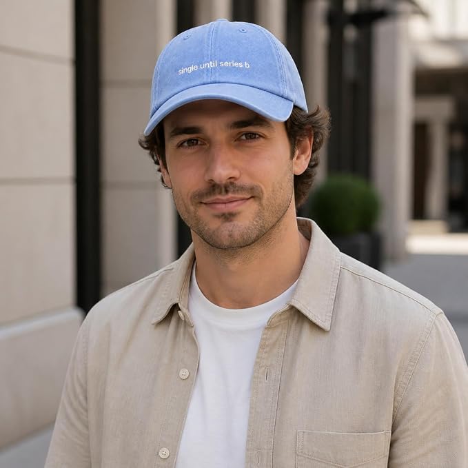 A man wearing a light blue baseball cap with white embroidered text reading 'single until series b' and a beige overshirt over a white t-shirt, photographed outdoors against a softly blurred building background.