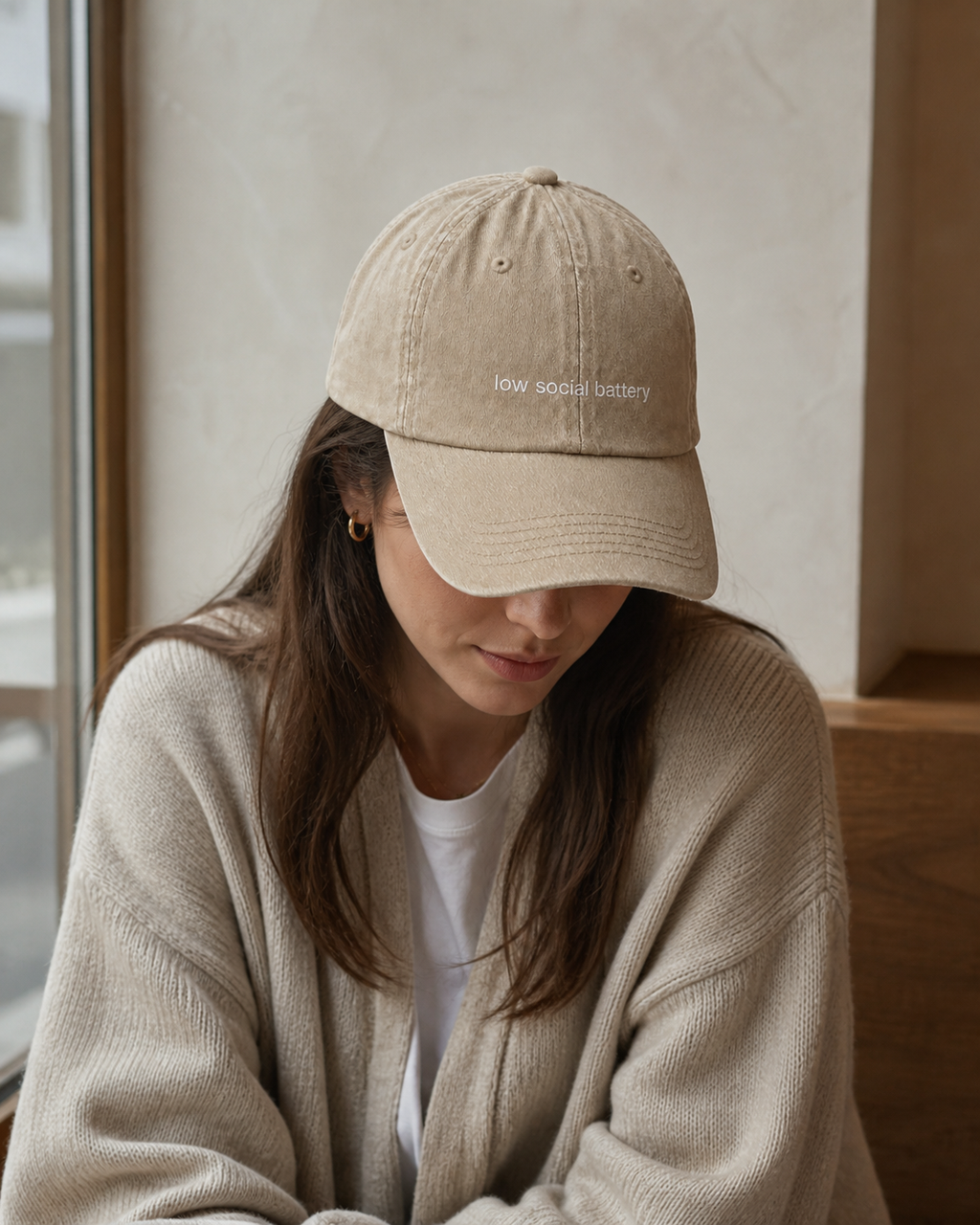 A person wearing a beige baseball cap embroidered with 'low social battery' in white, seated in a cafe.