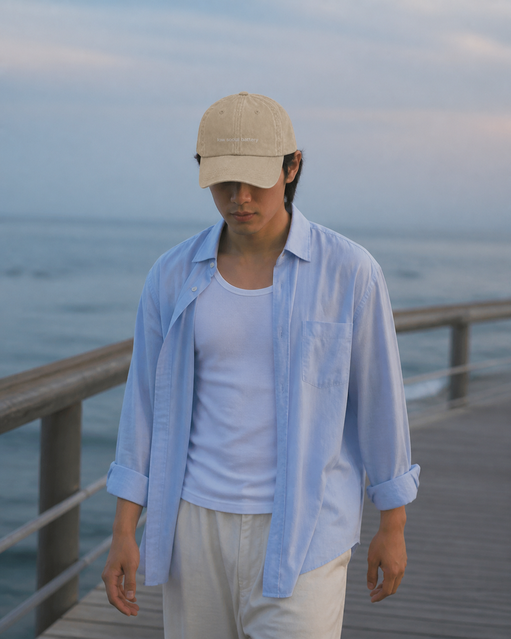 A person wearing a beige baseball cap embroidered with 'low social battery' in white by the sea.