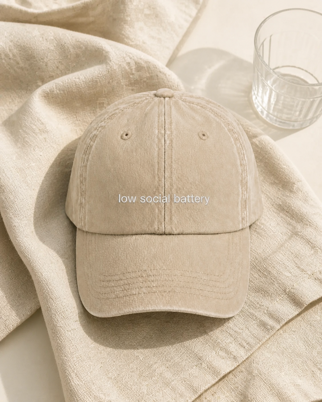 A beige baseball cap embroidered with 'low social battery' in white, laid flat on draped linen.