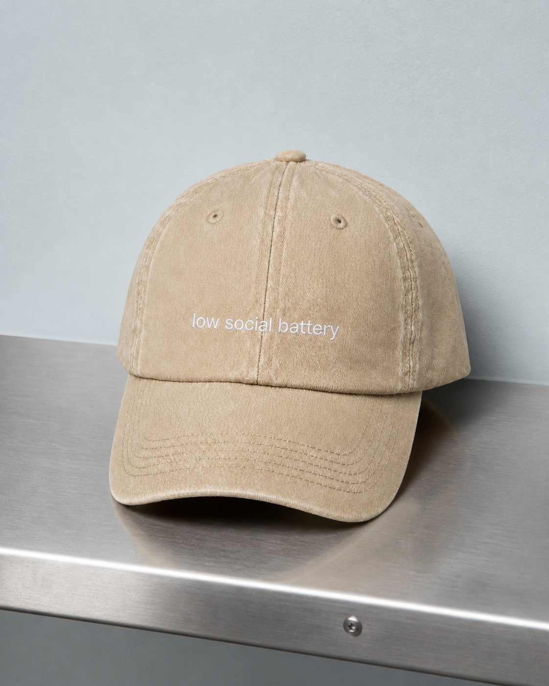A beige baseball cap embroidered with 'low social battery' in white, photographed on a brushed steel surface.