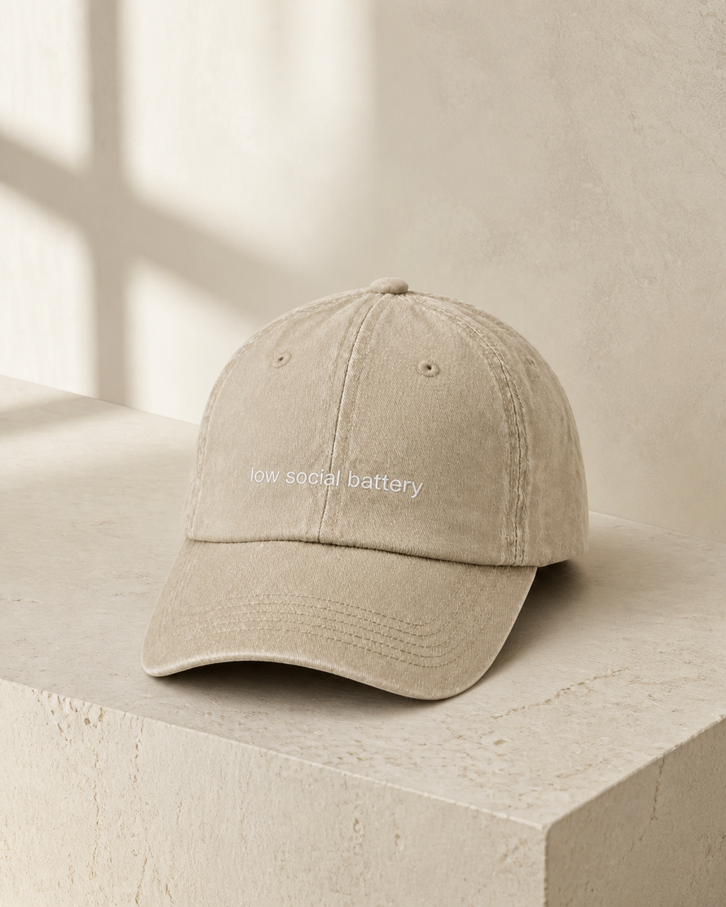 A beige baseball cap embroidered with 'low social battery' in white, displayed against a warm studio background.