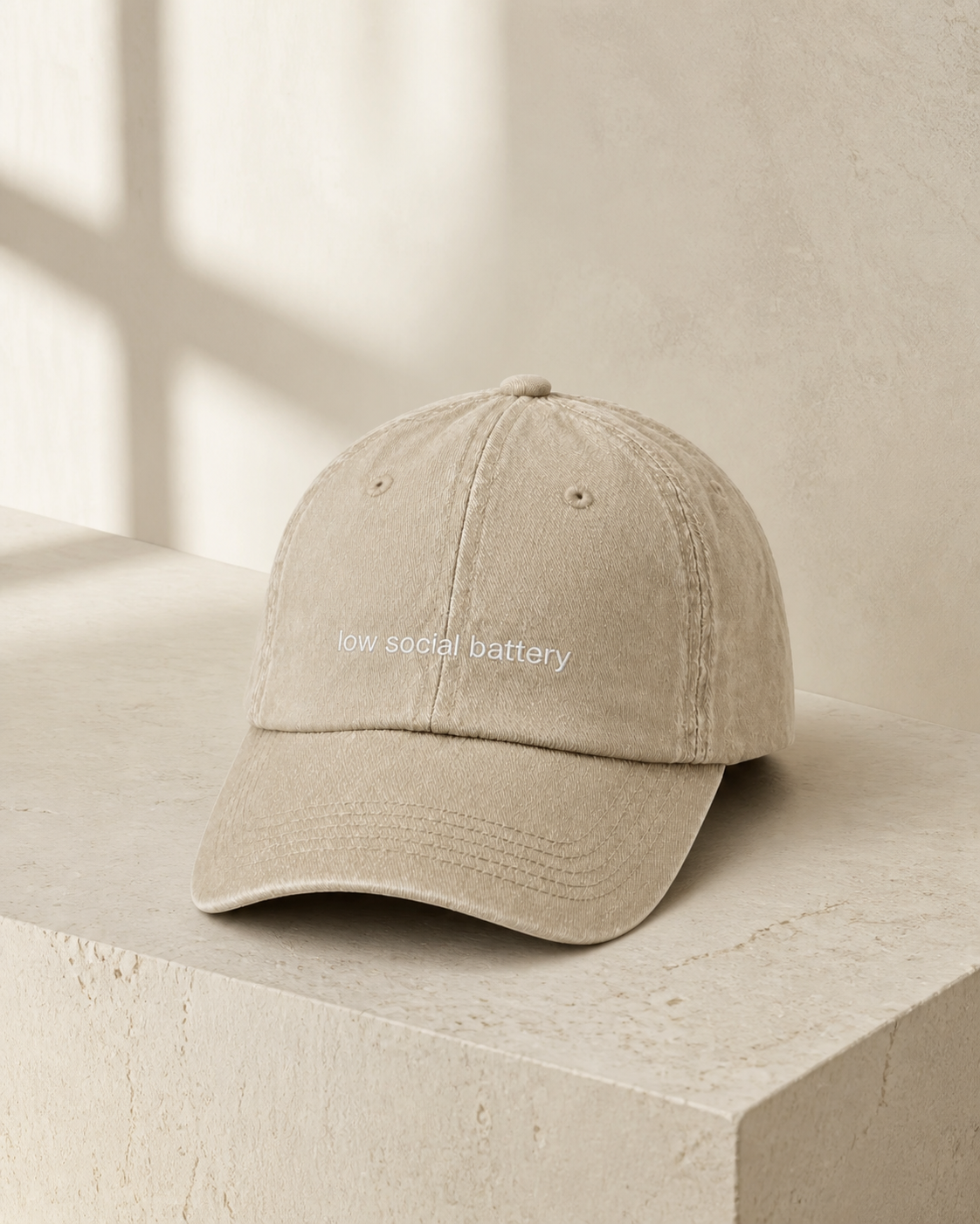 A beige baseball cap embroidered with 'low social battery' in white, displayed against a warm studio background.
