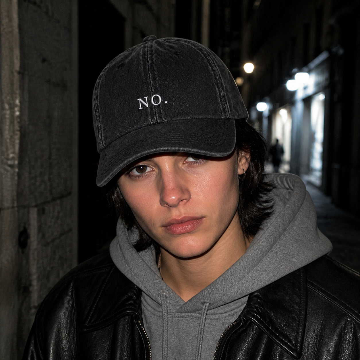 A person in a grey hoodie and black leather jacket wearing the washed black 'NO.' cap on a narrow street at night, lit by flash.