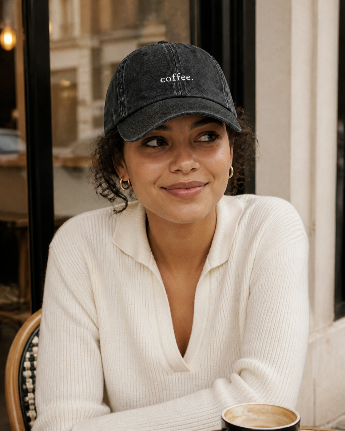 A woman in a cream knit top wearing the washed black 'coffee.' cap at a cafe window with a latte in front of her.