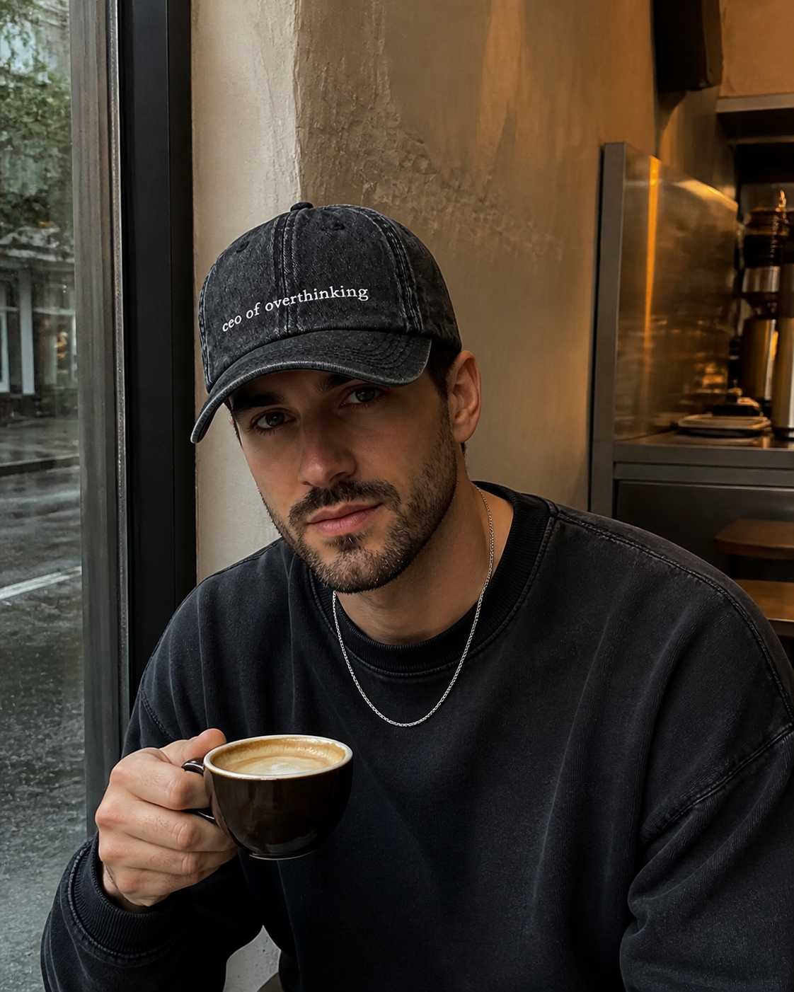 A man in a black sweatshirt wearing the washed black 'ceo of overthinking' cap and holding a cappuccino by a rainy cafe window.