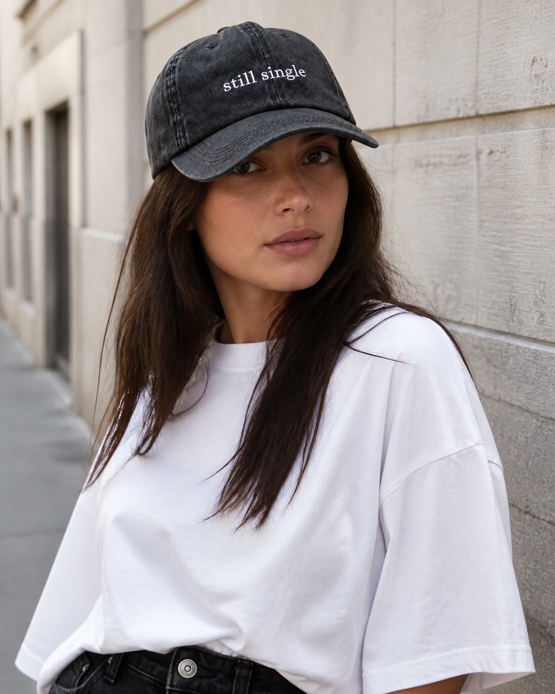 A person wearing a washed black baseball cap embroidered with 'still single' in white on a city street.