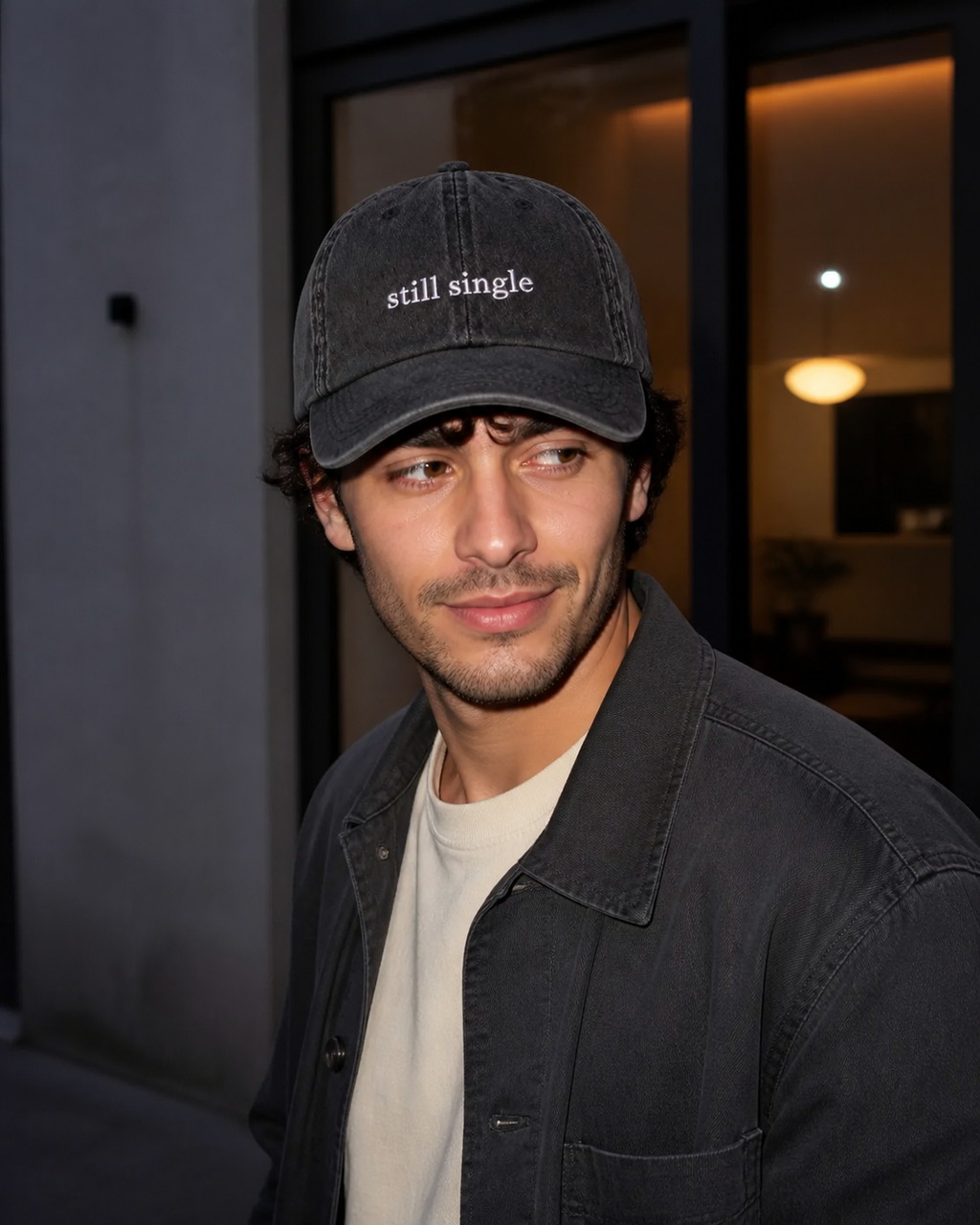 A person wearing a washed black baseball cap embroidered with 'still single' in white, photographed outdoors in golden-hour light.