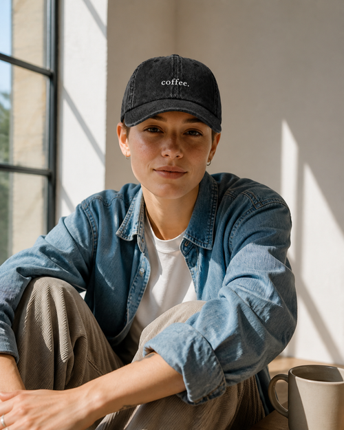 A woman in a denim shirt wearing the washed black 'coffee.' cap, seated by a sunlit loft window beside a ceramic mug.