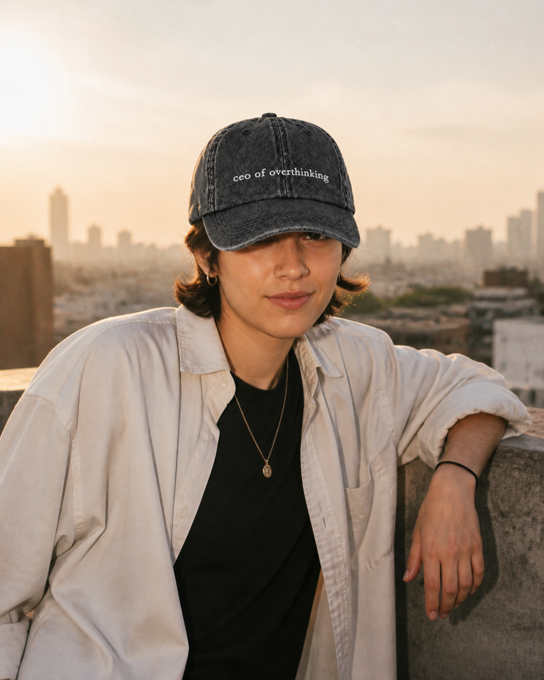 A person in a cream overshirt wearing the washed black 'ceo of overthinking' cap on a rooftop with a city skyline at sunset.