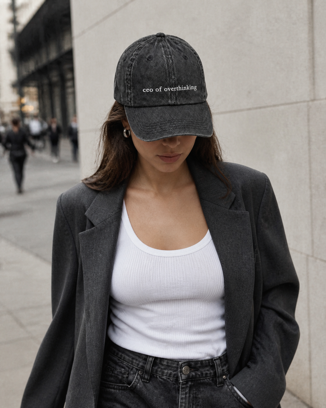 A woman in a grey blazer and white tank top wearing the washed black 'ceo of overthinking' cap pulled low on a city street.