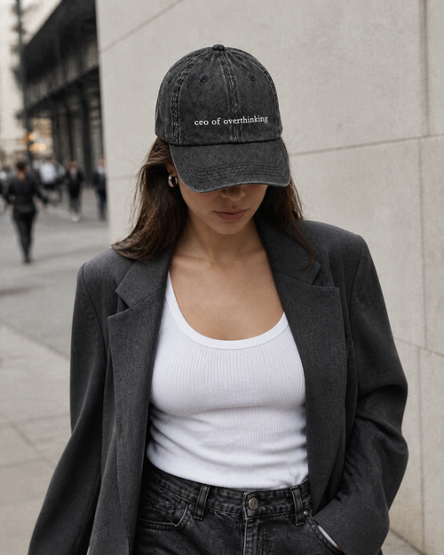 A woman in a grey blazer and white tank top wearing the washed black 'ceo of overthinking' cap pulled low on a city street.