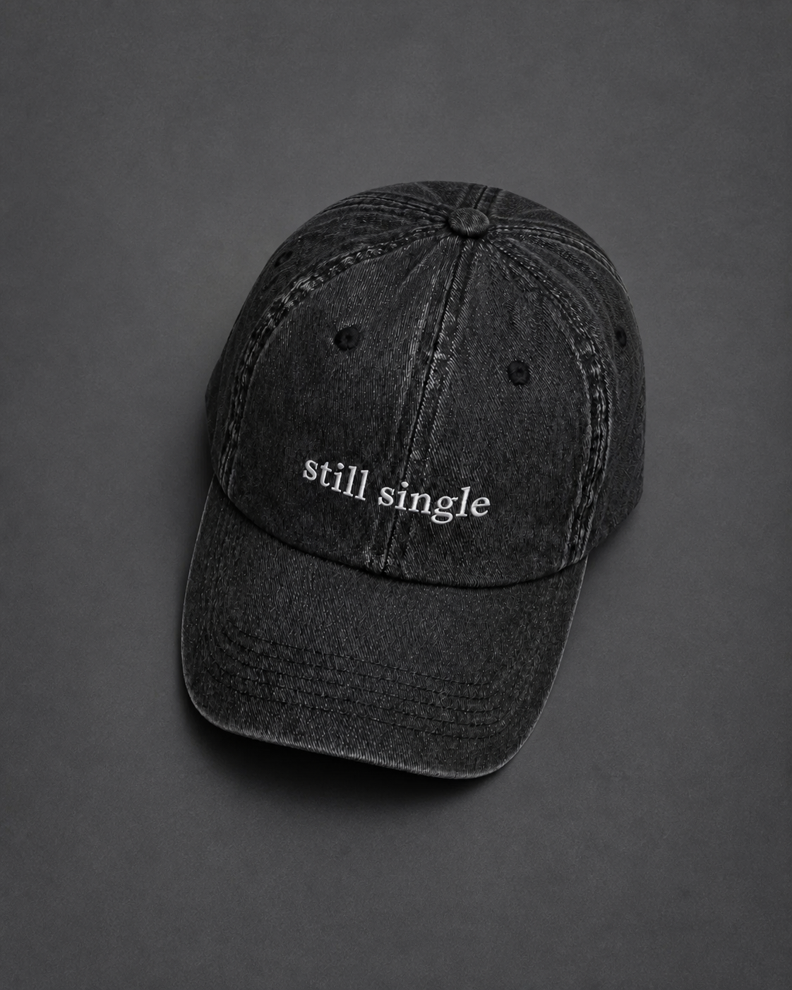 A washed black baseball cap embroidered with 'still single' in white, photographed on a charcoal surface.