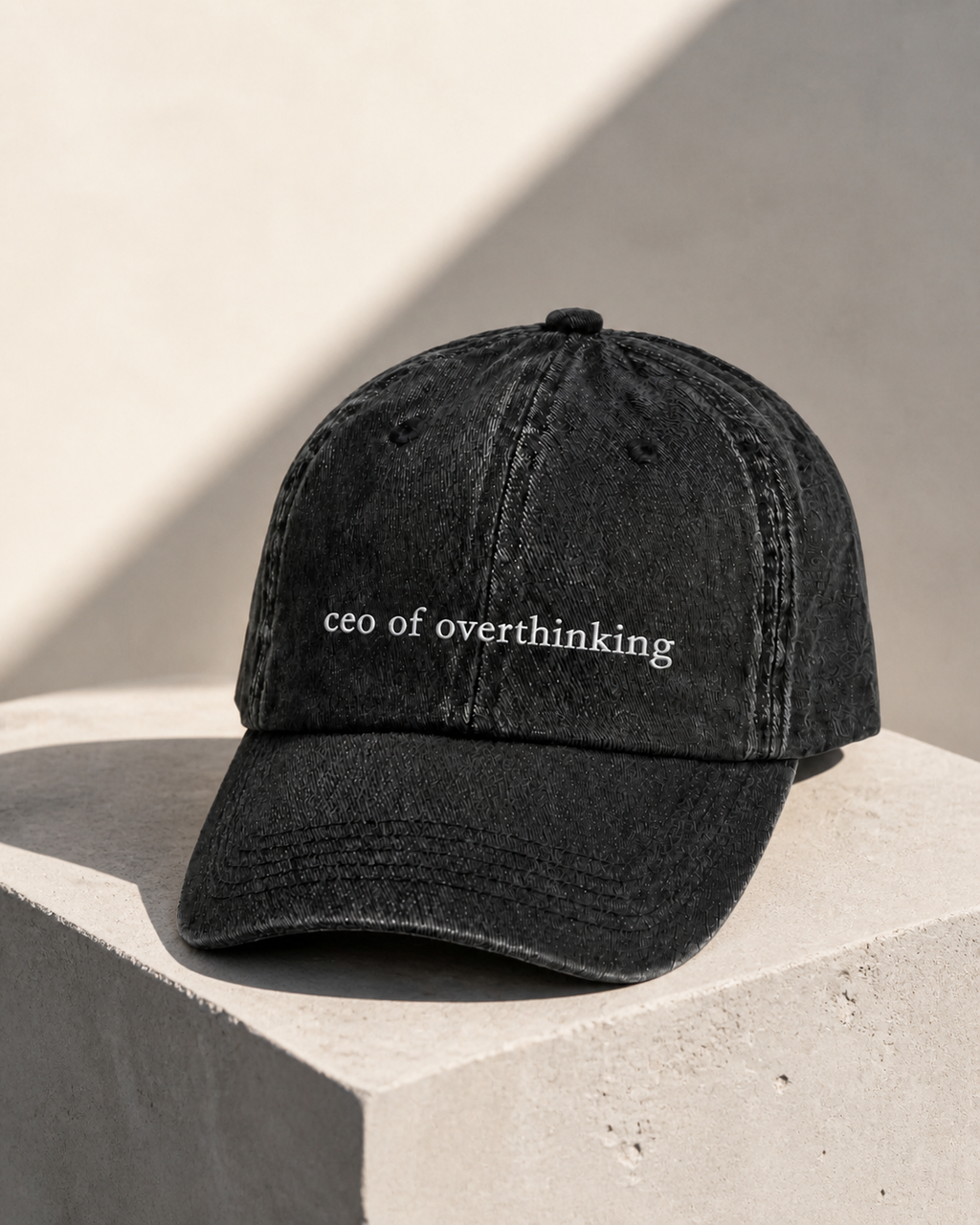 The washed black 'ceo of overthinking' cap resting on a concrete block in hard afternoon sunlight.