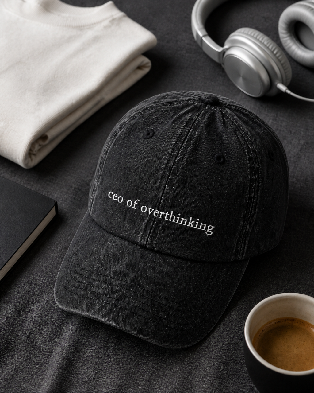 The washed black 'ceo of overthinking' cap in a flat lay with a folded cream sweatshirt, silver headphones, a notebook and an espresso.