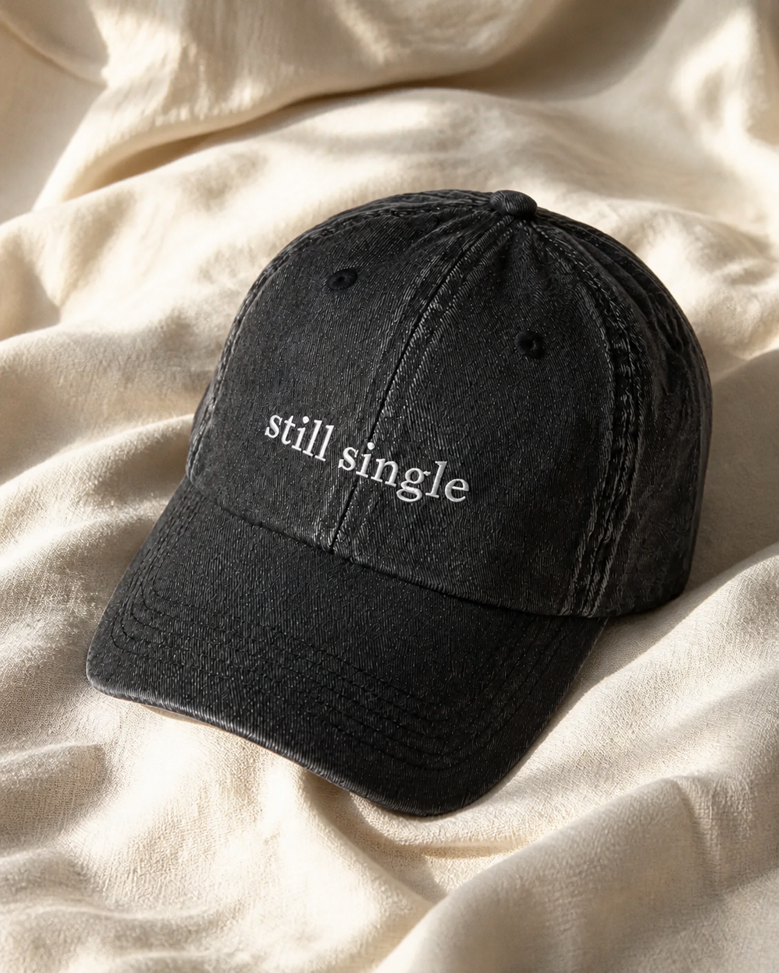 A washed black baseball cap embroidered with 'still single' in white, resting on draped linen.
