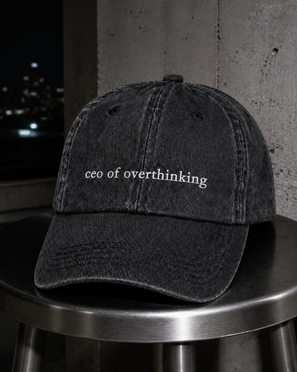The washed black 'ceo of overthinking' cap on a metal stool at night, lit by flash against a concrete wall.