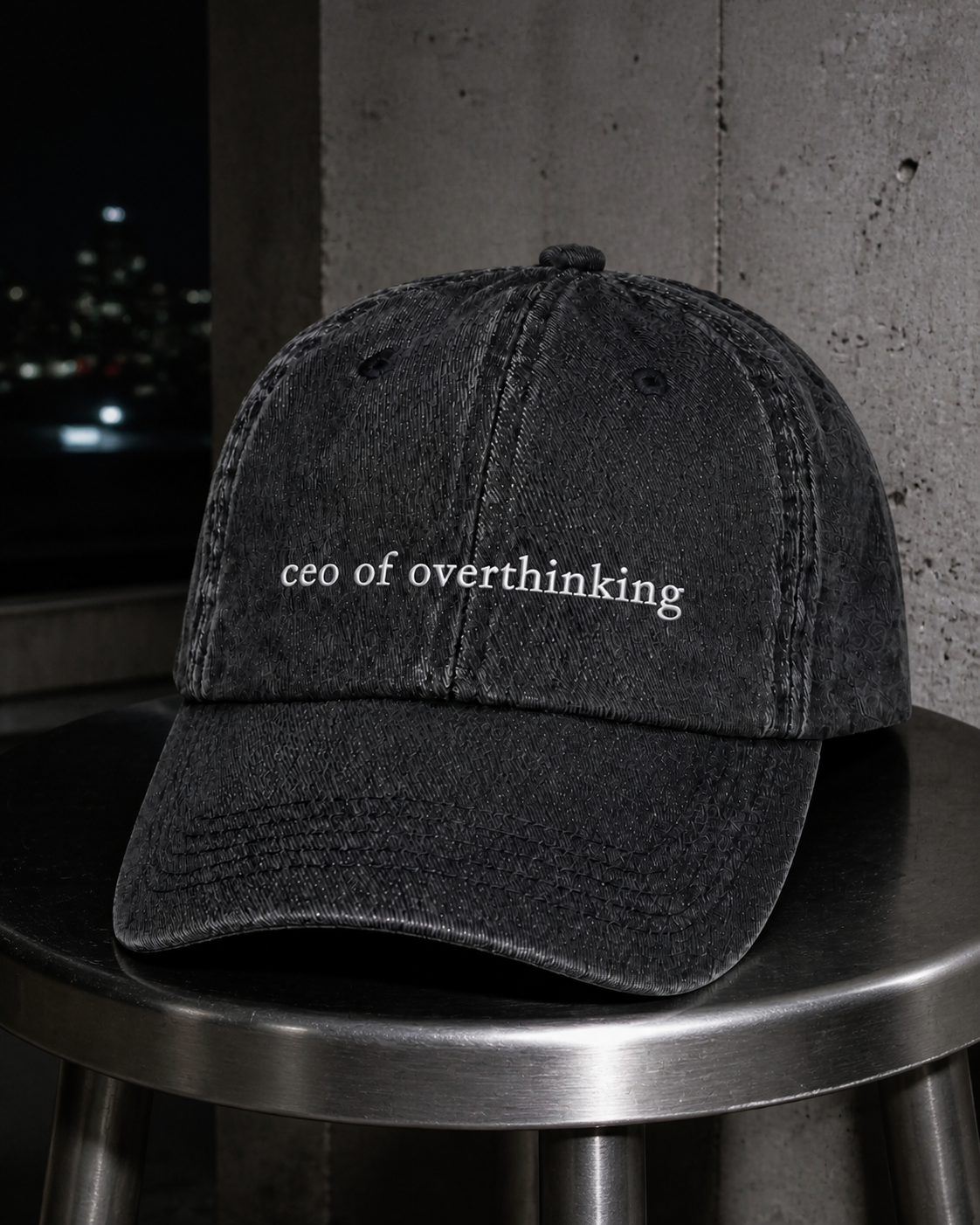 The washed black 'ceo of overthinking' cap on a metal stool at night, lit by flash against a concrete wall.