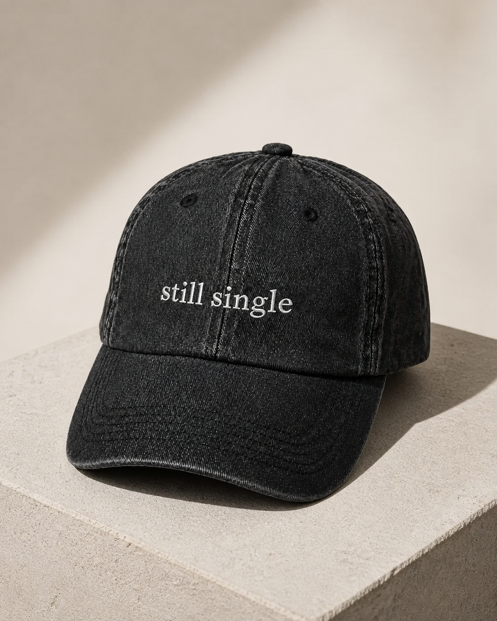 A washed black baseball cap embroidered with 'still single' in white, displayed on a stone plinth.