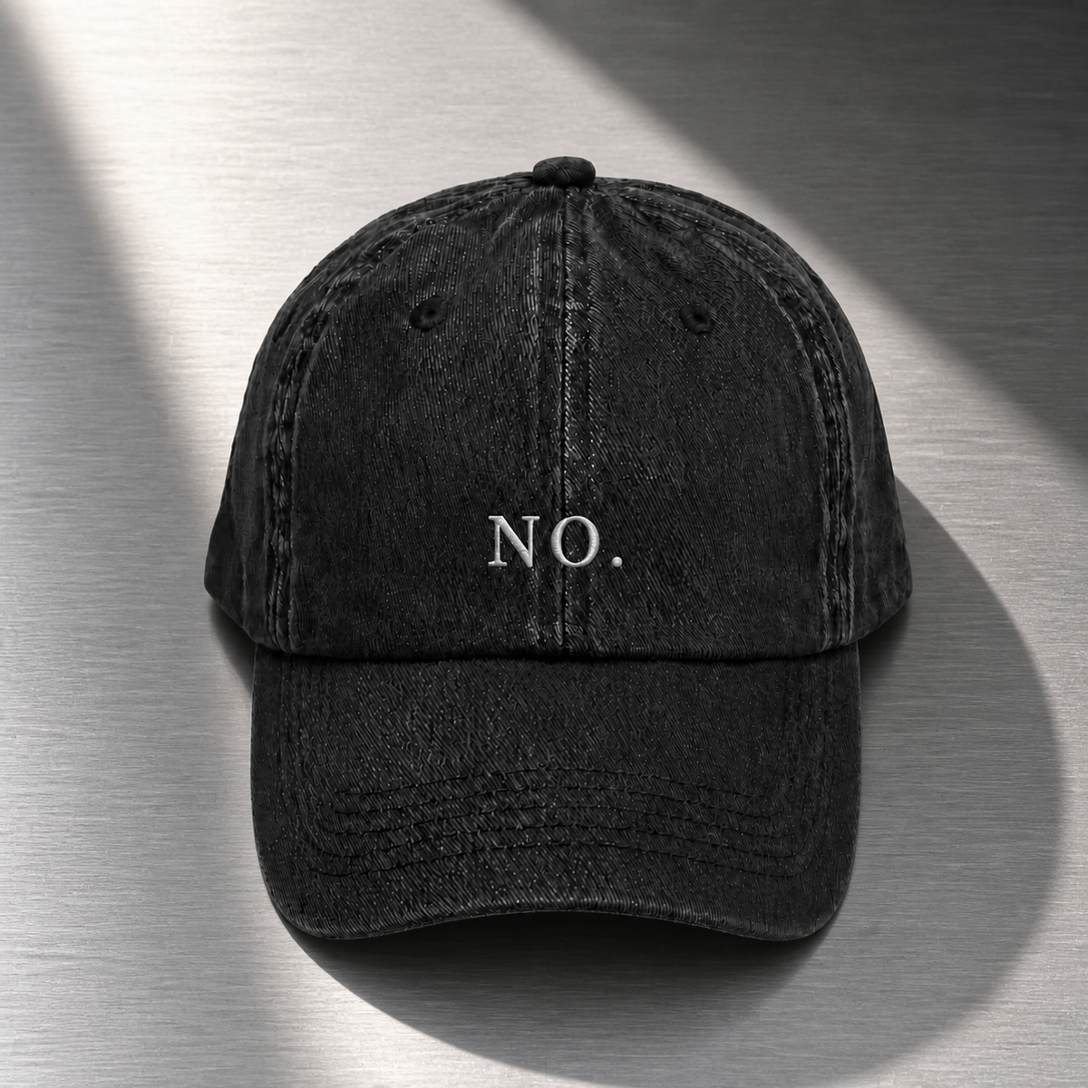 The washed black 'NO.' cap laid flat on brushed steel with a diagonal shaft of light across it.
