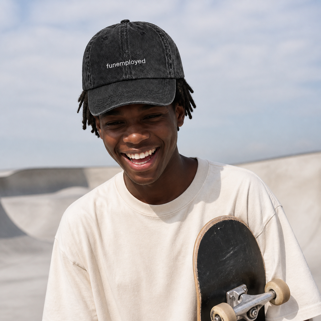 A smiling young man at a skatepark wearing a faded black baseball cap with the text 'funemployed' and a cream-colored t-shirt while holding a skateboard.