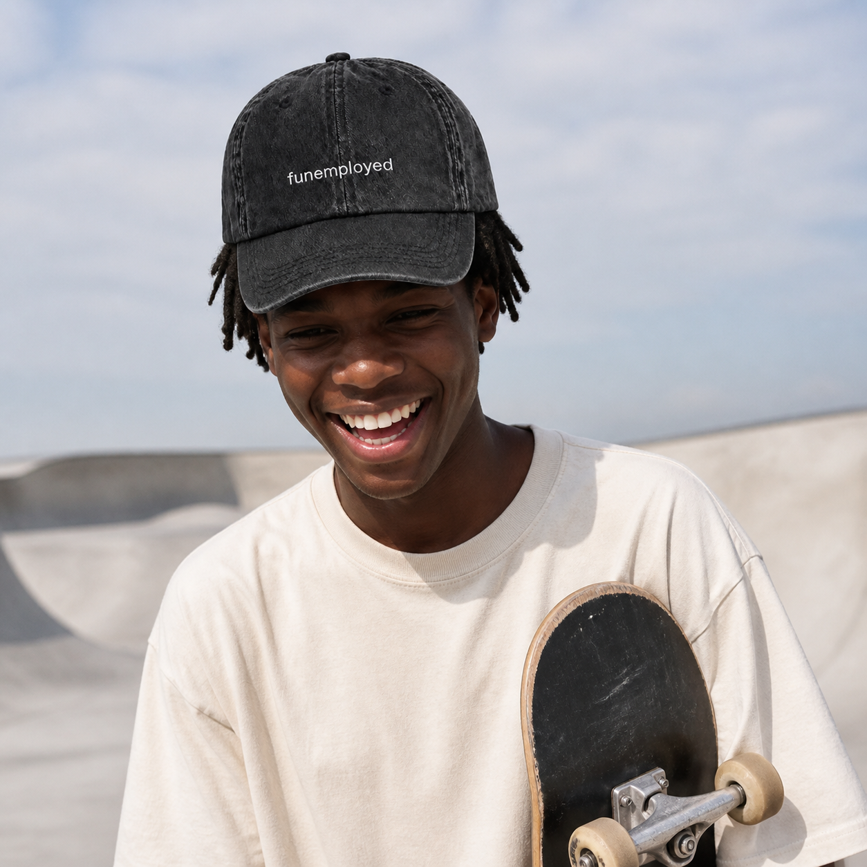 A smiling young man at a skatepark wearing a faded black baseball cap with the text 'funemployed' and a cream-colored t-shirt while holding a skateboard.