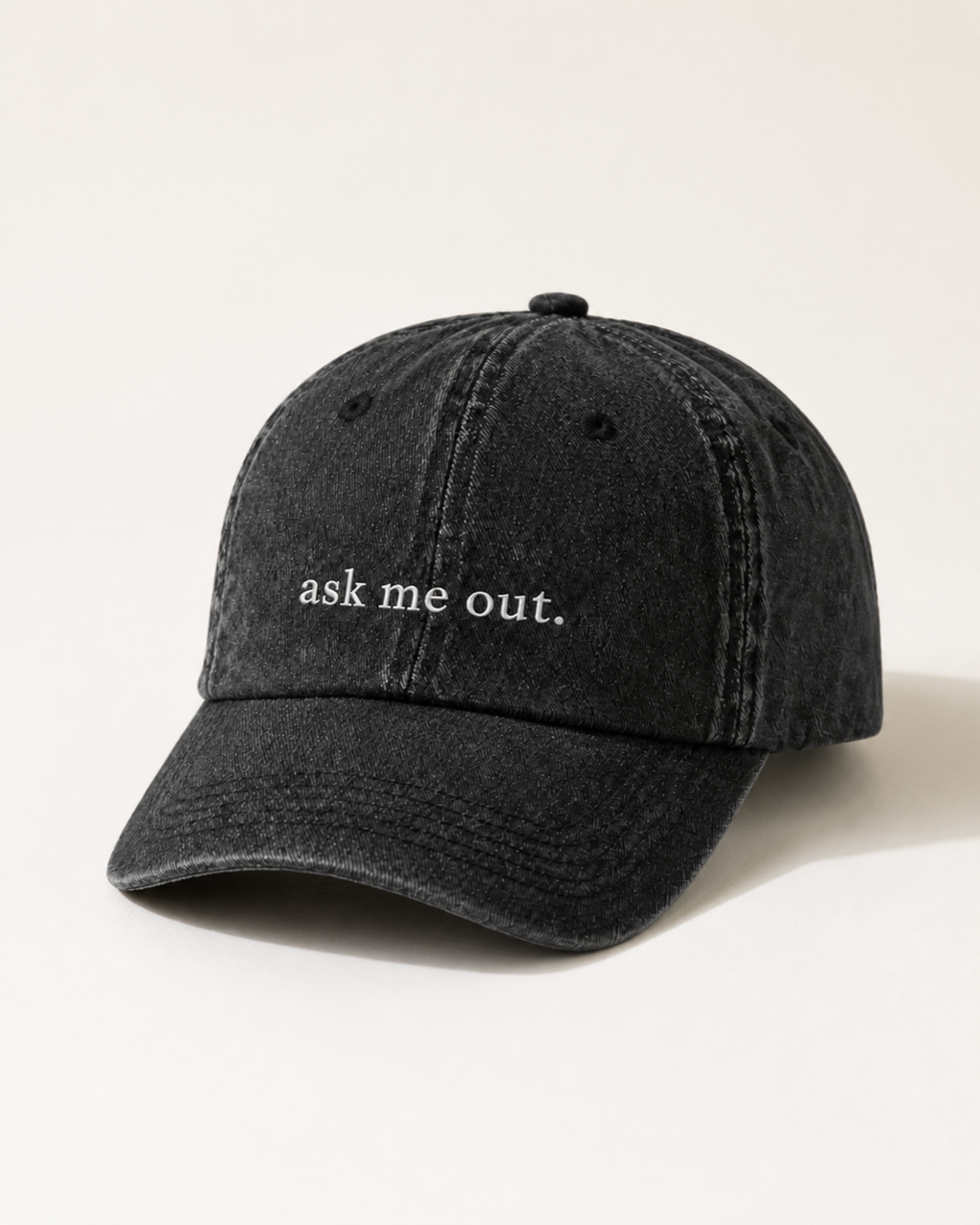 Washed black 'ask me out.' cap photographed at a three-quarter angle against a warm cream studio backdrop.
