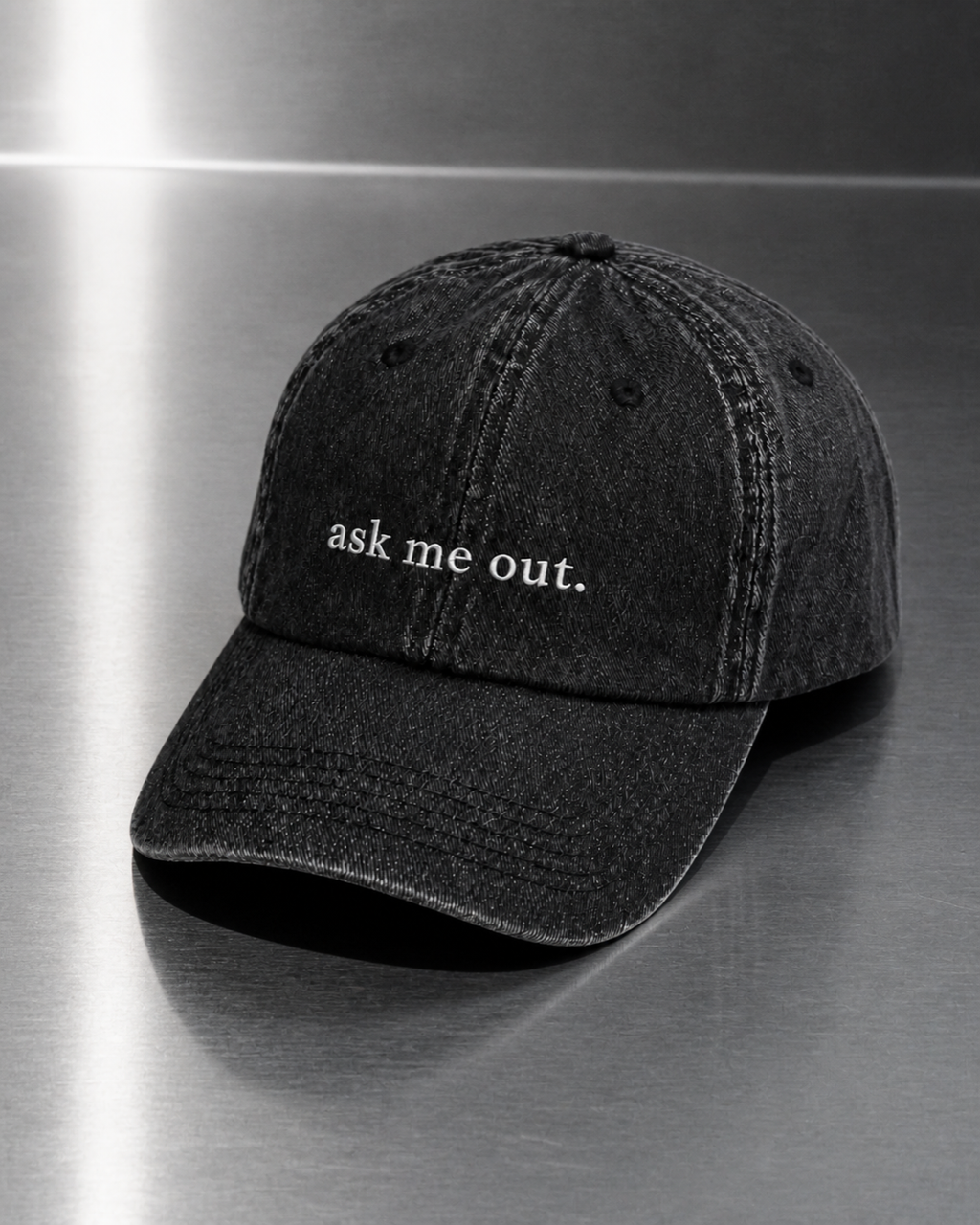 Washed black 'ask me out.' cap resting on a brushed steel surface under cool directional light.