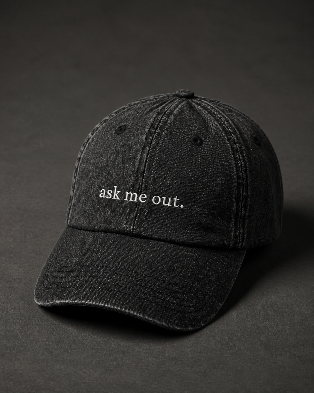 Washed black 'ask me out.' cap on a dark charcoal backdrop, lit from the side to pick out the embroidery.