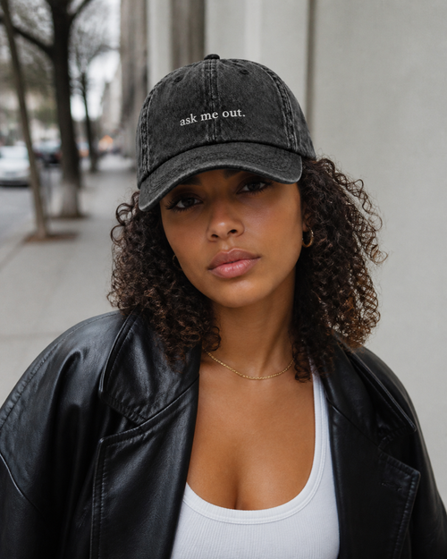 A woman in a black leather jacket and white tank top wearing the washed black 'ask me out.' cap on a city street.
