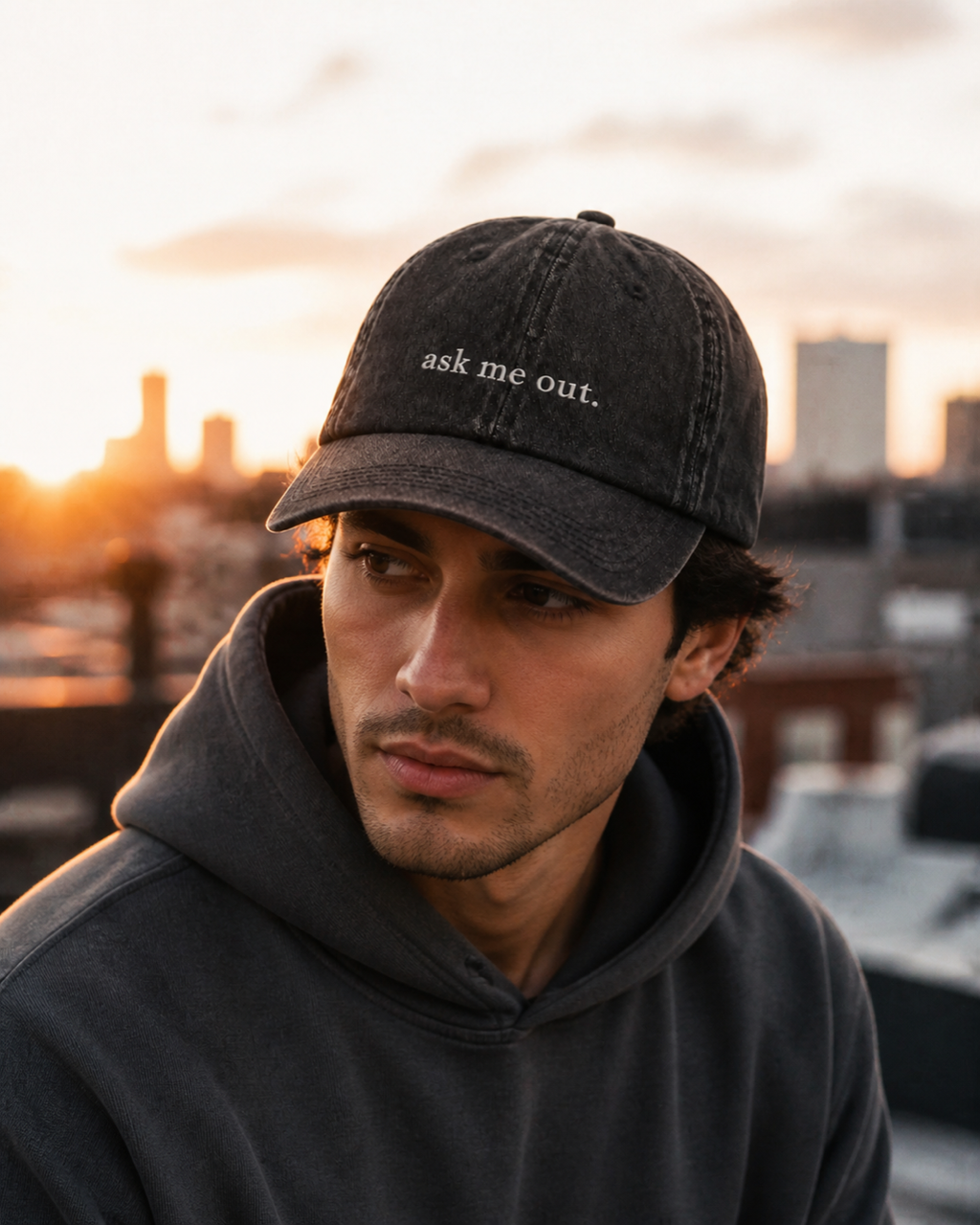 A man in a dark hooded sweatshirt wearing the washed black 'ask me out.' cap on a rooftop at sunset, city skyline behind him.
