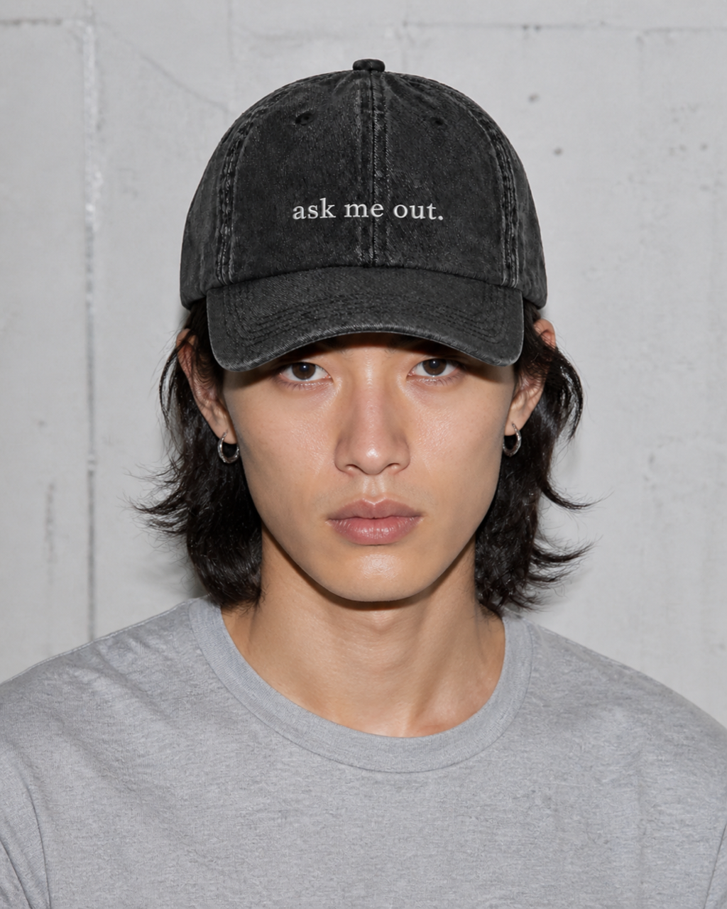 A person in a grey t-shirt wearing the washed black 'ask me out.' cap against a bare concrete wall.