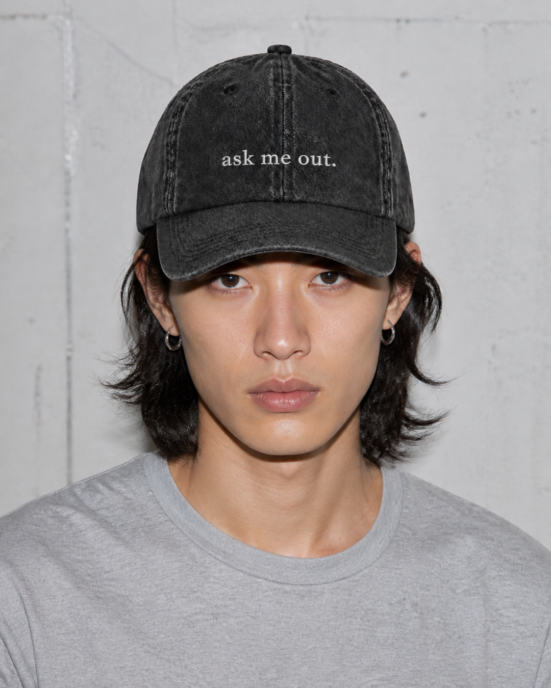 A person in a grey t-shirt wearing the washed black 'ask me out.' cap against a bare concrete wall.