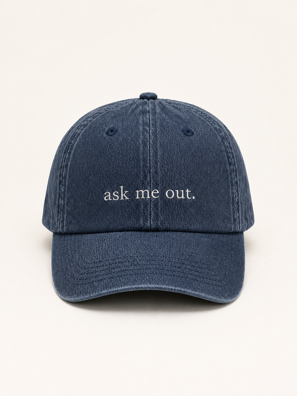 Washed navy 'ask me out.' cap photographed face-on against a warm cream studio backdrop.