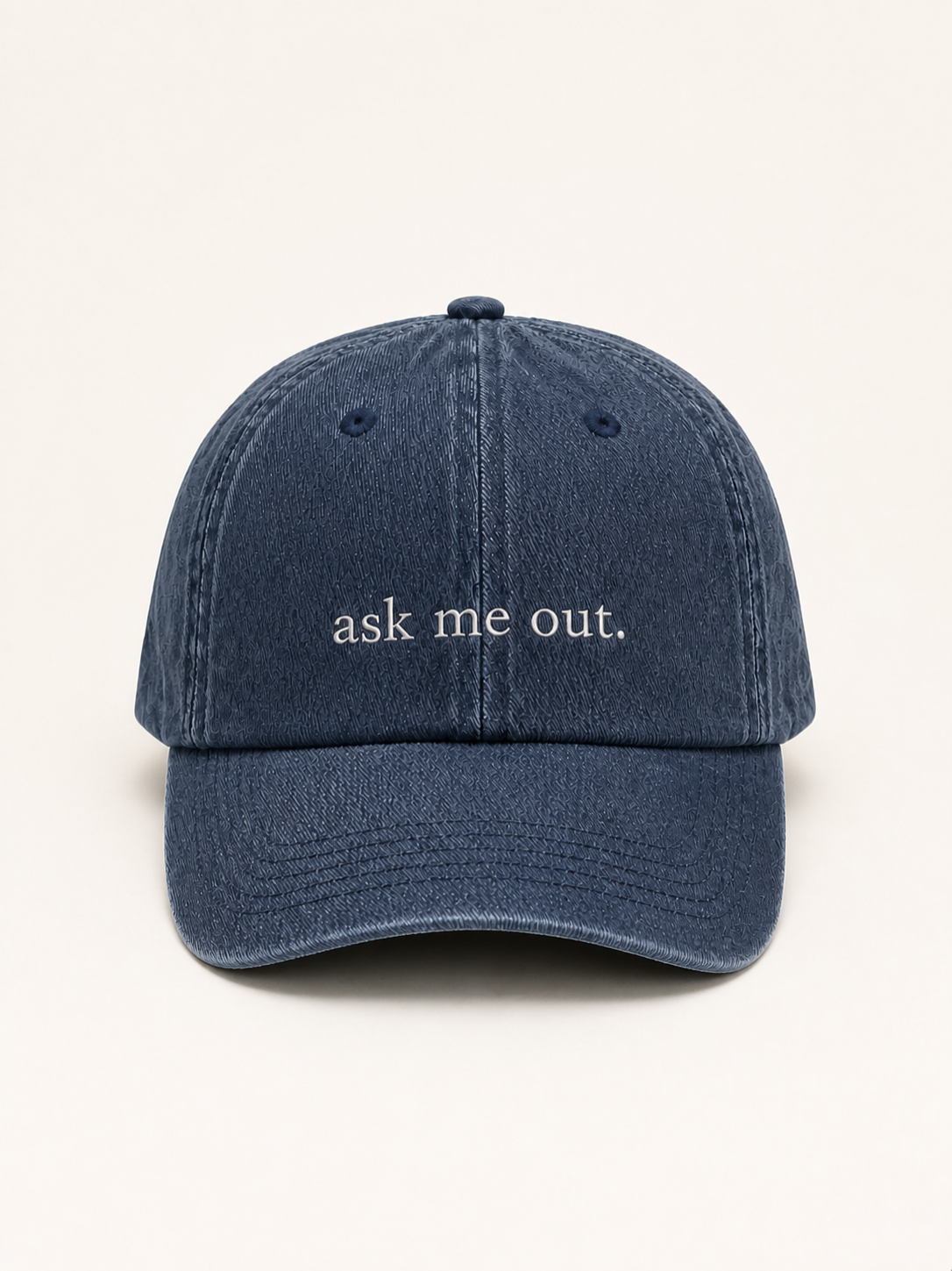 Washed navy 'ask me out.' cap photographed face-on against a warm cream studio backdrop.
