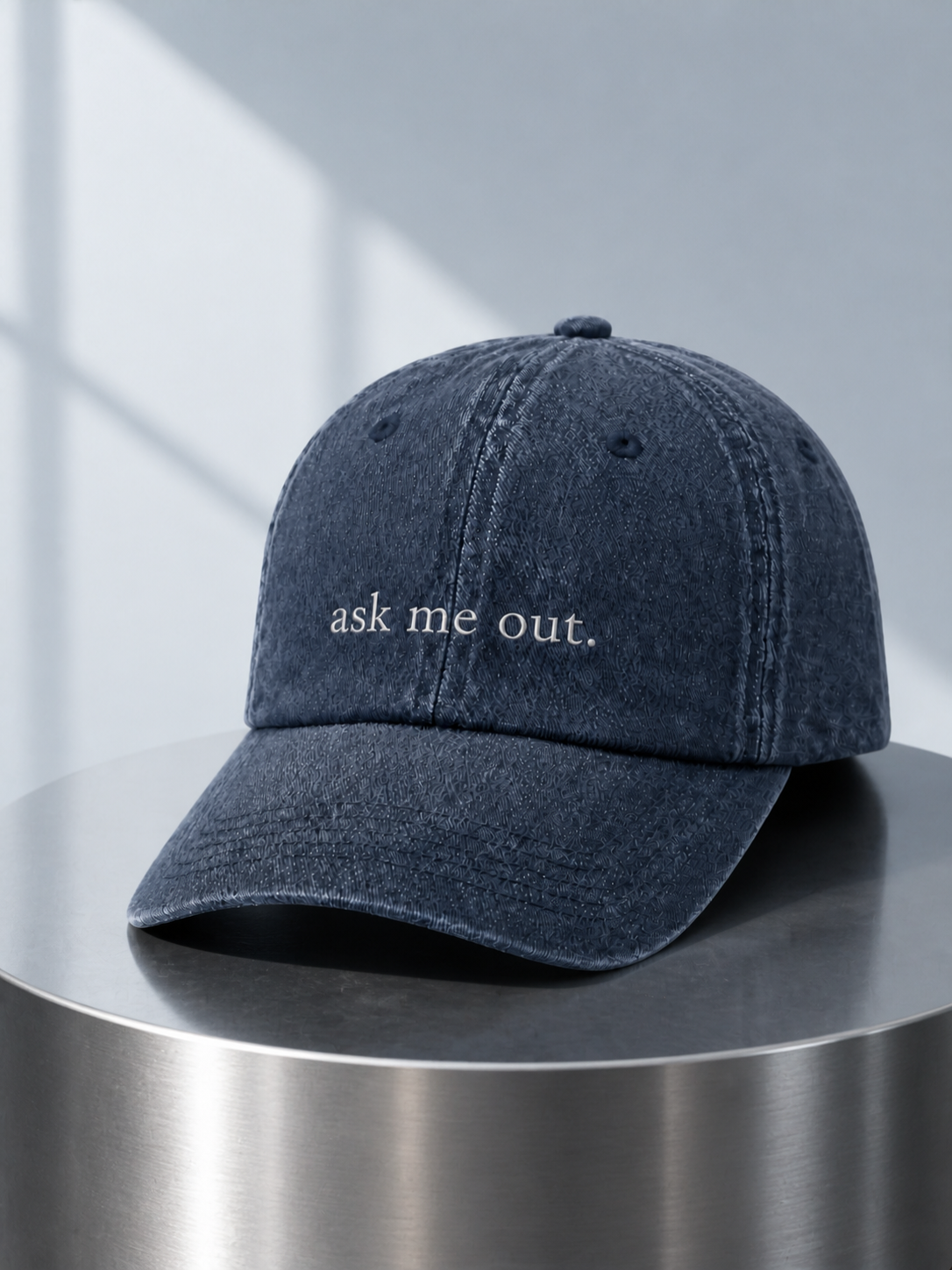 Washed navy 'ask me out.' cap displayed on a polished steel cylinder plinth in cool daylight.